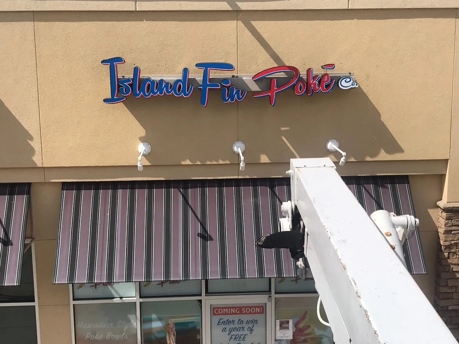Island Fin Poké Co. red and blue channel letters being installed from a lift above a striped awning