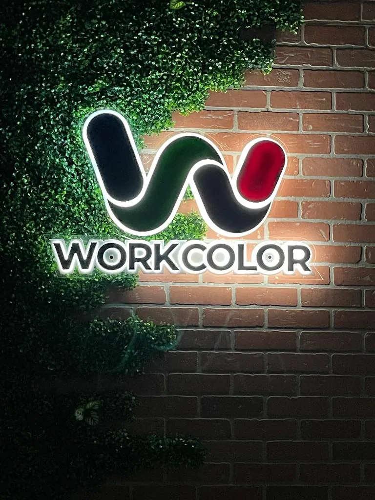 Illuminated Work Color logo sign on a brick wall framed by greenery