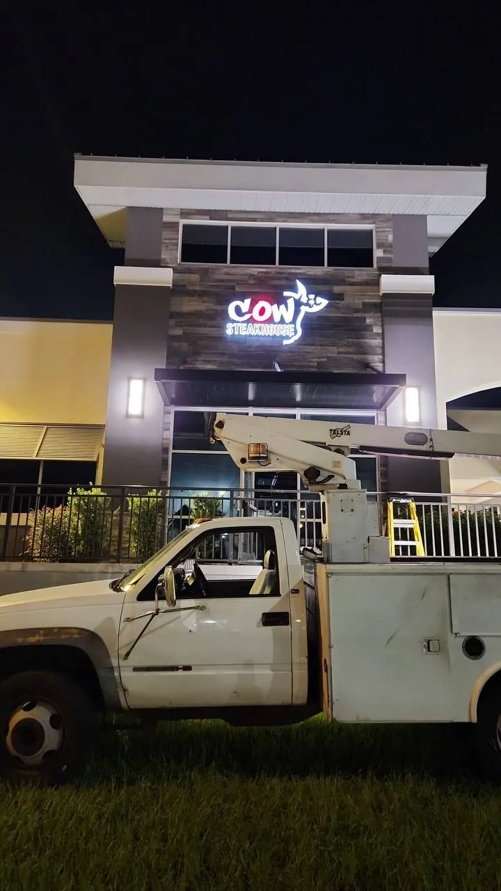Bucket truck parked below the newly lit Cow Steakhouse sign on a restaurant facade at night