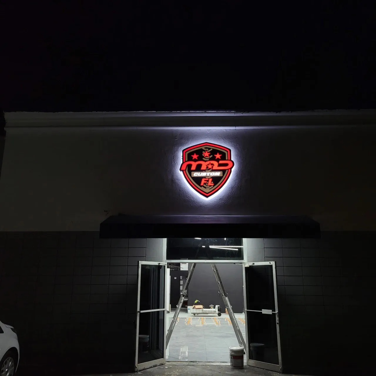 MAD Custom FL shield logo sign with a white halo glow above a shop entrance at night