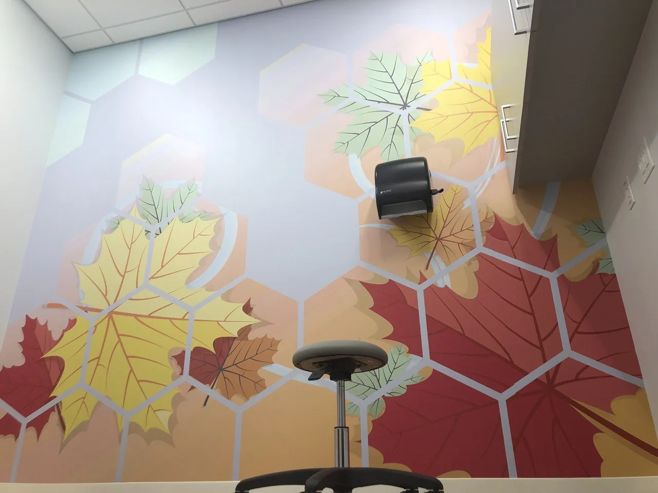 Corner room wrapped in a red, yellow and green maple-leaf hexagon mural with a stool