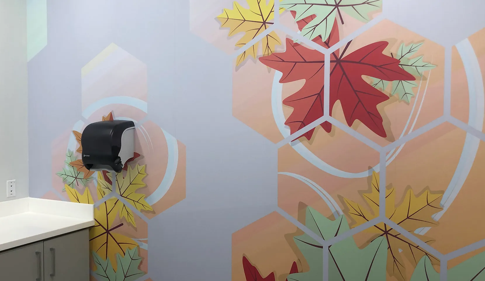 Wall wrap specs: the finished autumn maple-leaf hexagon mural beside a counter and paper-towel dispenser